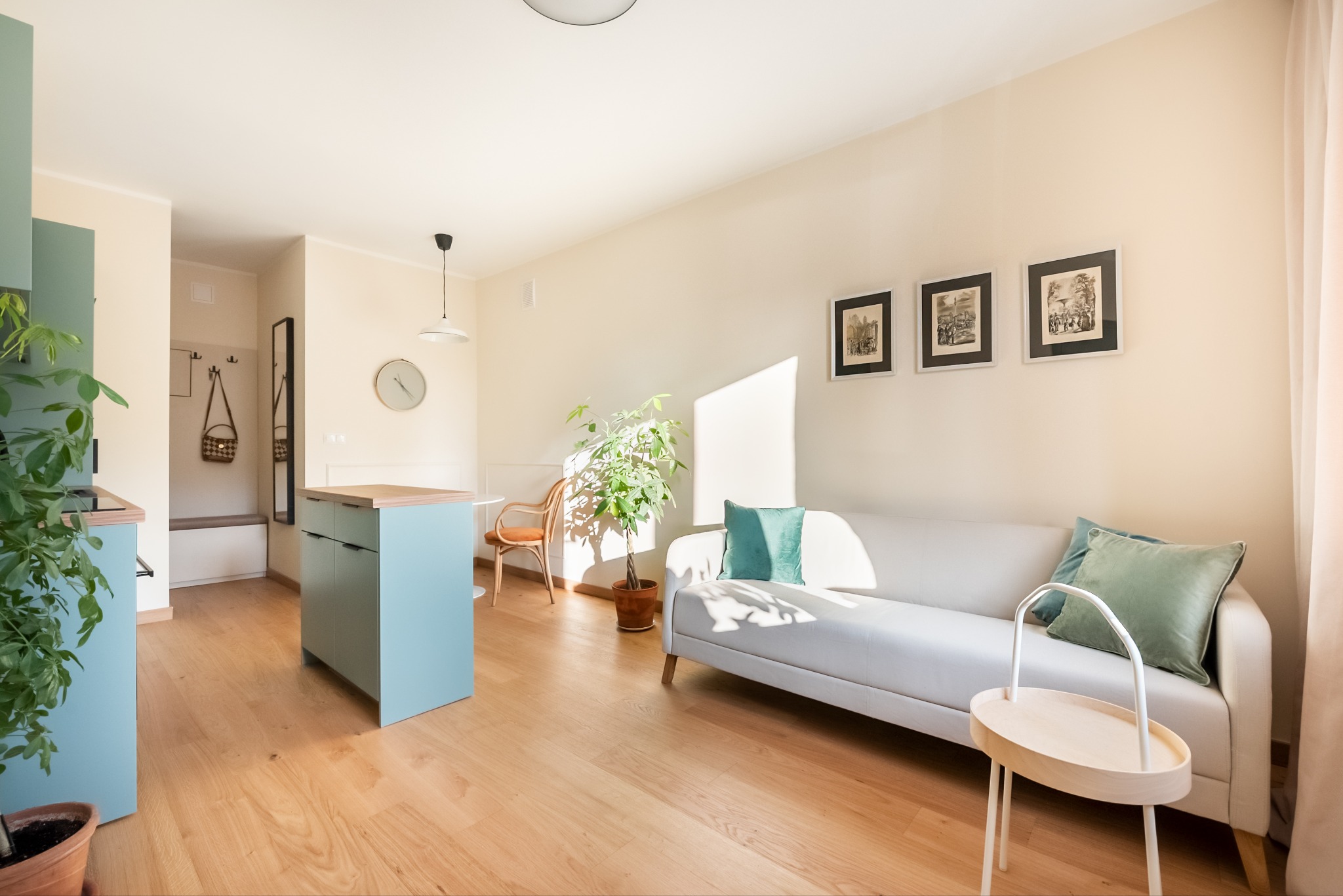 Jasne wnętrze mieszkania z drewnianą podłogą, turkusową wyspą kuchenną, białym narożnikiem z zielonymi poduszkami i roślinami doniczkowymi.