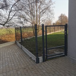 Drenaż Ogrodzenia Marek Chmielewski - Nowoczesne, antracytowe ogrodzenie panelowe z bramką, zamontowane na betonowej podmurówce, widoczne z perspektywy brukowanego podjazdu, z trawnikiem i drzewami w tle.