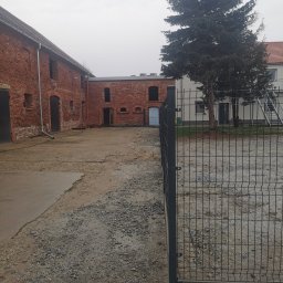 Drenaż Ogrodzenia Marek Chmielewski - Nowoczesne, metalowe ogrodzenie panelowe na żwirowym podłożu, w tle ceglany budynek gospodarczy i wysoka choinka w pochmurny dzień.