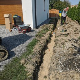 Drenaż Ogrodzenia Marek Chmielewski - Pracownik kopie wąski wykop w ziemi obok domu, widoczne rury kanalizacyjne, taczka, opony i narzędzia