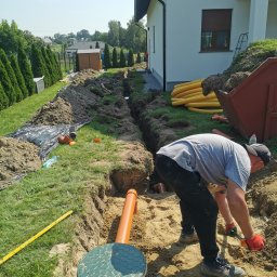 Drenaż Ogrodzenia Marek Chmielewski - Pracownik w czerwonych rękawicach kopie łopatą w wykopie pod drenaż rozsączający, widoczna studzienka drenażowa z zieloną pokrywą, rury drenażowe, dom jednorodzinny w tle.