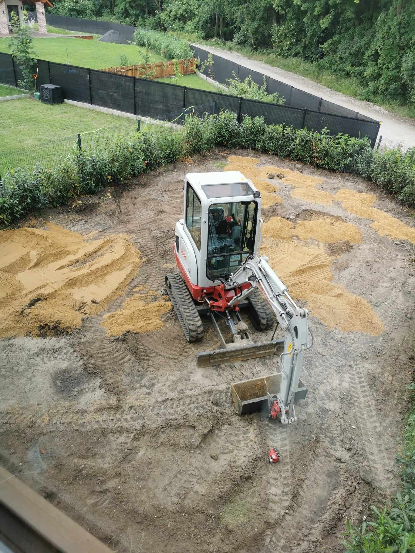 Widok z góry na mini-koparkę na gąsienicach w trakcie prac ziemnych, z rozplantowanym piaskiem na przygotowanym terenie pod budowę, w tle widoczne ogrodzenie panelowe i zieleń.