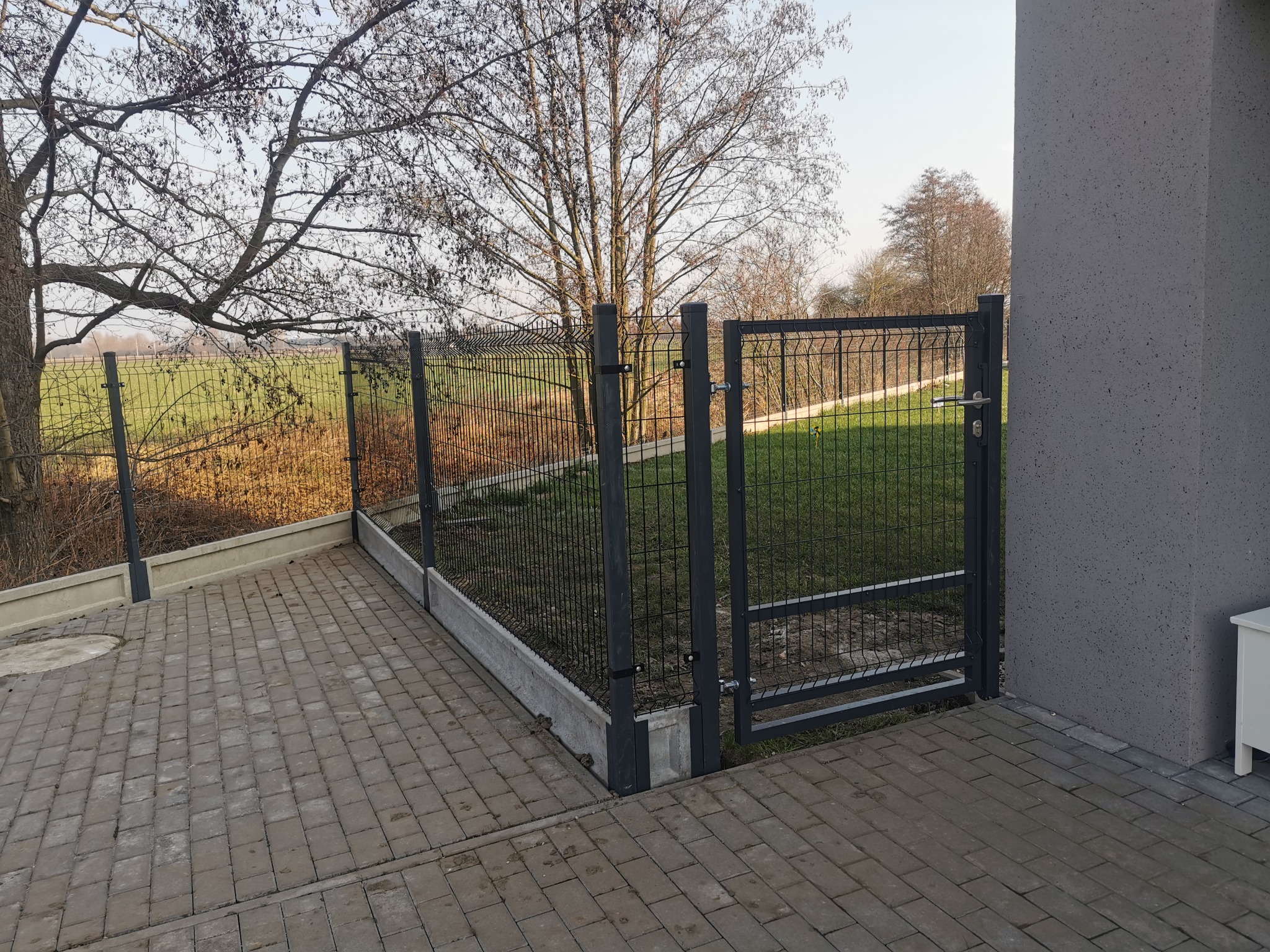 Nowoczesne, antracytowe ogrodzenie panelowe z bramką, zamontowane na betonowej podmurówce, widoczne z perspektywy brukowanego podjazdu, z trawnikiem i drzewami w tle.