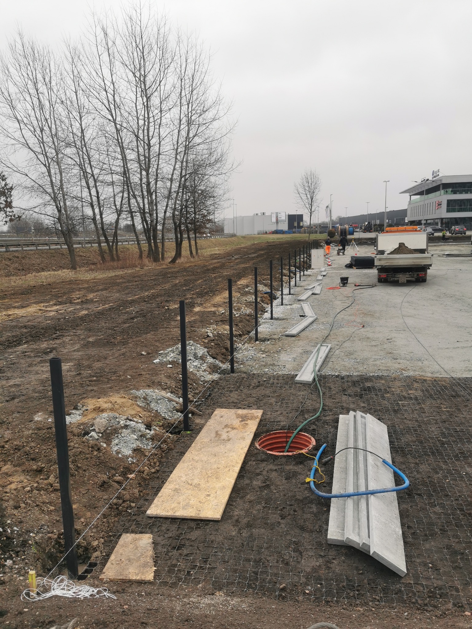 Przygotowanie terenu pod budowę ogrodzenia: wbite słupki, rozciągnięty sznurek wyznaczający linię, ułożone krawężniki, widoczny właz kanalizacyjny i ciężarówka z piaskiem w tle.