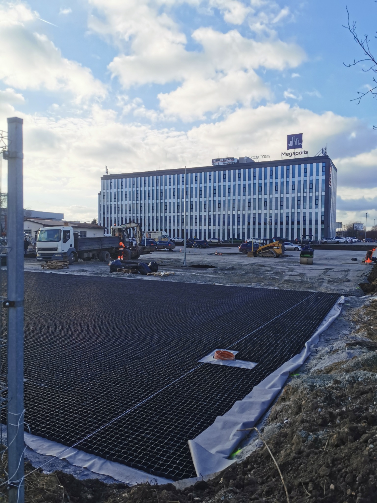 Przygotowanie terenu pod parking: widok na rozłożoną geokratę na podłożu, w tle budynek biurowy Megapolis, ciężarówka i maszyny budowlane.