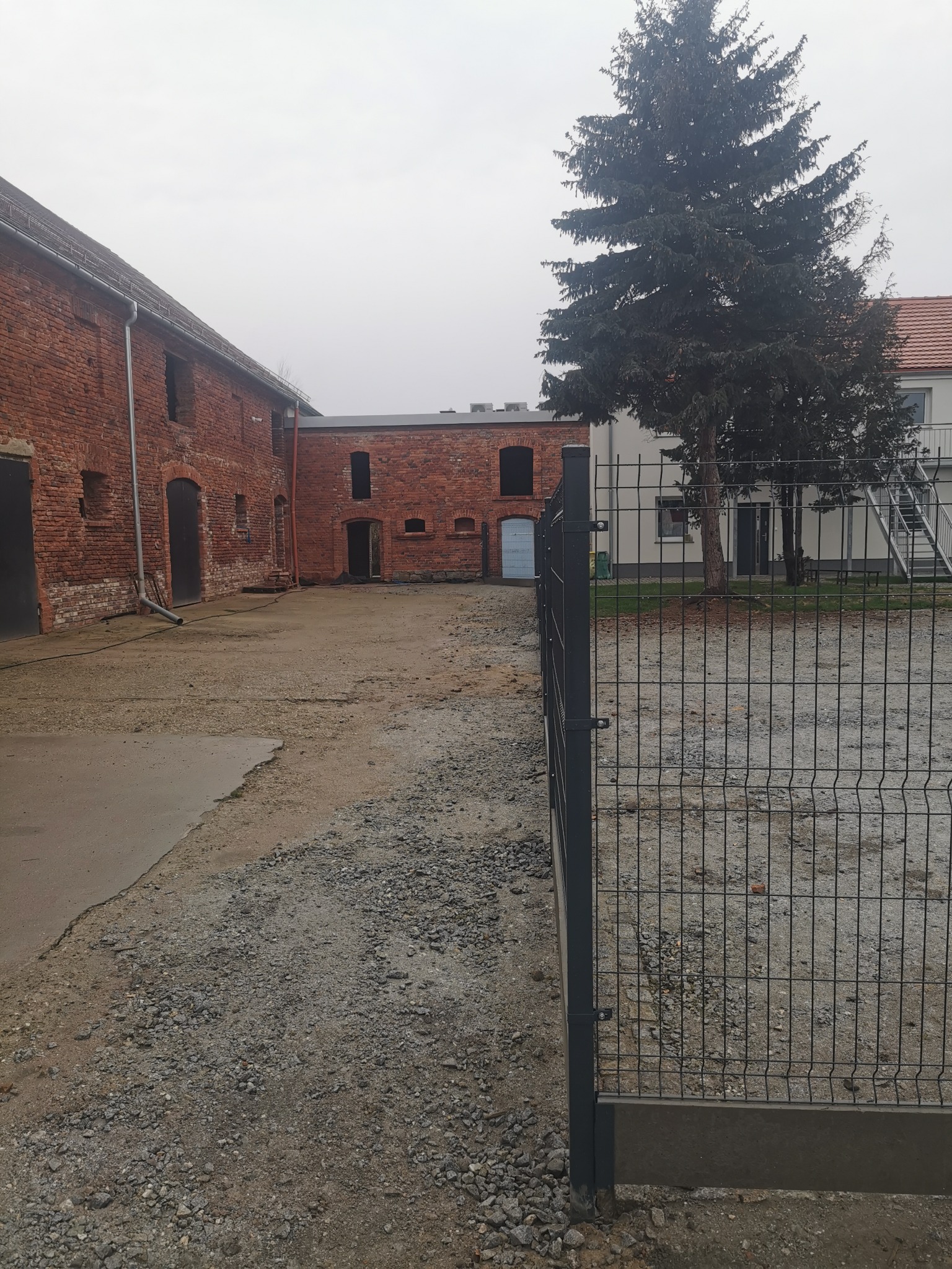 Nowoczesne, metalowe ogrodzenie panelowe na żwirowym podłożu, w tle ceglany budynek gospodarczy i wysoka choinka w pochmurny dzień.