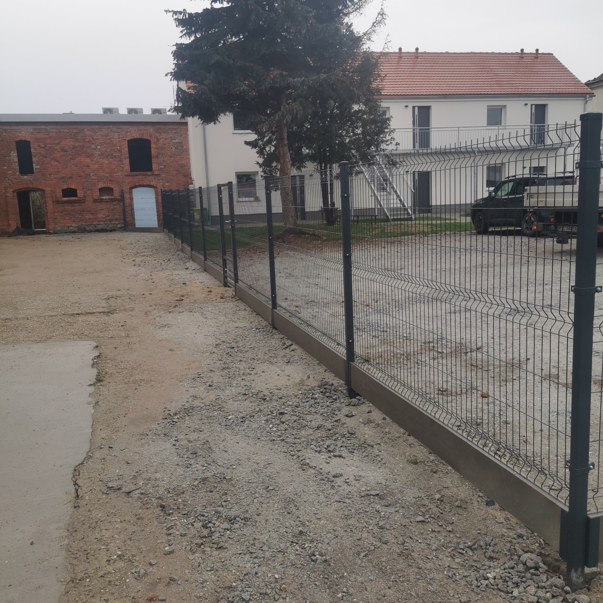 Nowoczesne ogrodzenie panelowe z podmurówką betonową na żwirowym podłożu, w tle budynki gospodarcze z cegły i nowy dom z balkonami, pochmurny dzień.