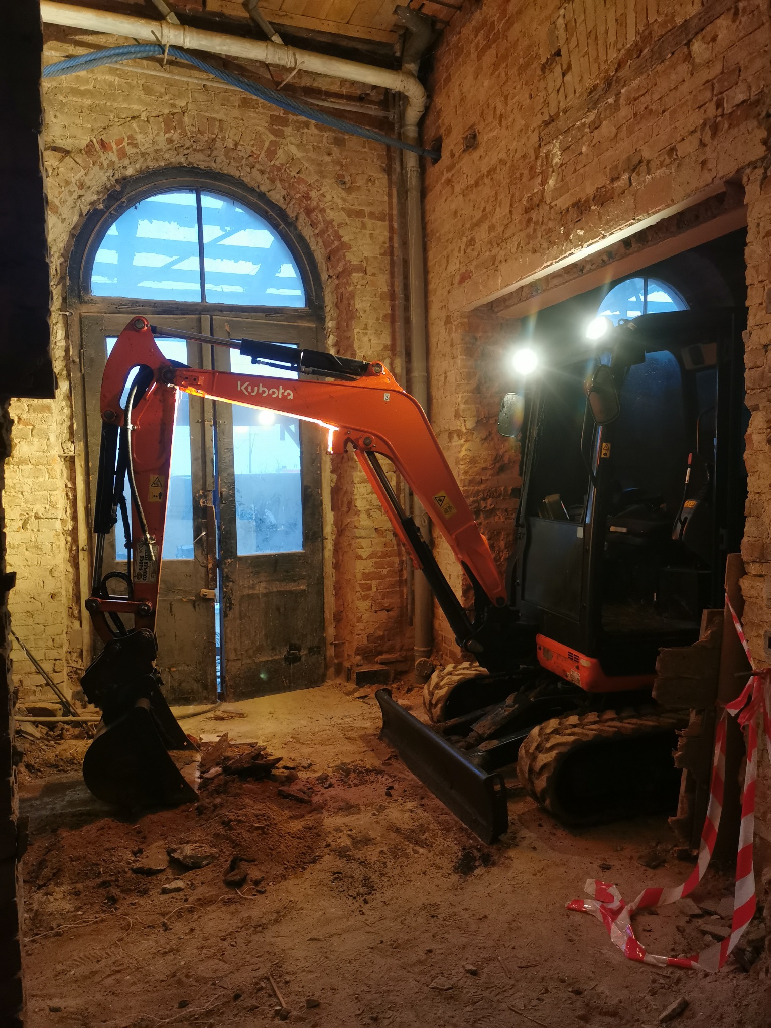Pomarańczowa mini koparka Kubota wewnątrz budynku z cegły, przygotowująca teren, widoczne stare, drewniane drzwi z łukowym oknem w tle, oświetlenie robocze koparki.