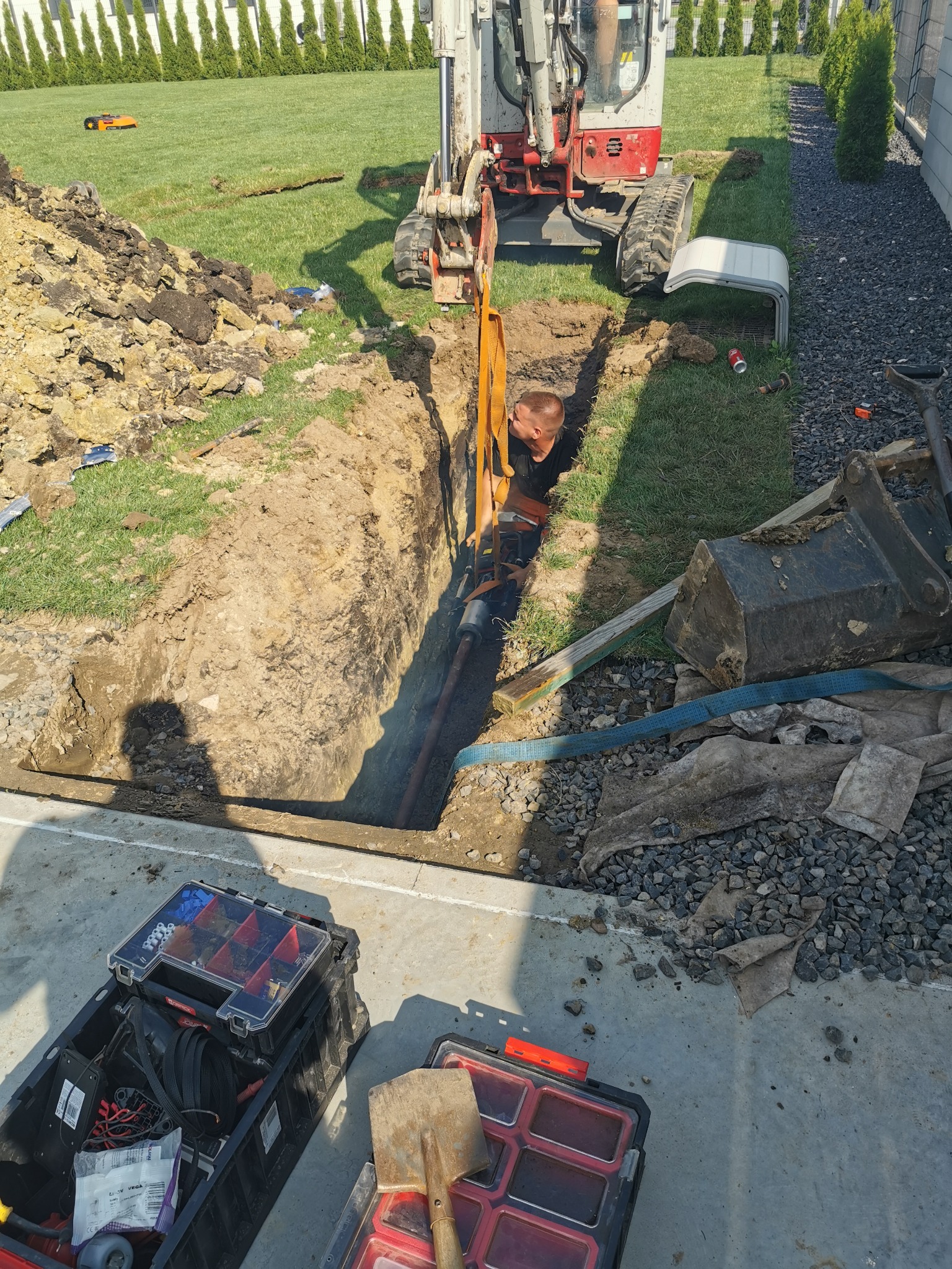 Pracownik w wykopie pod instalacje, widoczna rura, mini koparka, skrzynki narzędziowe, trawnik z tujami w tle.