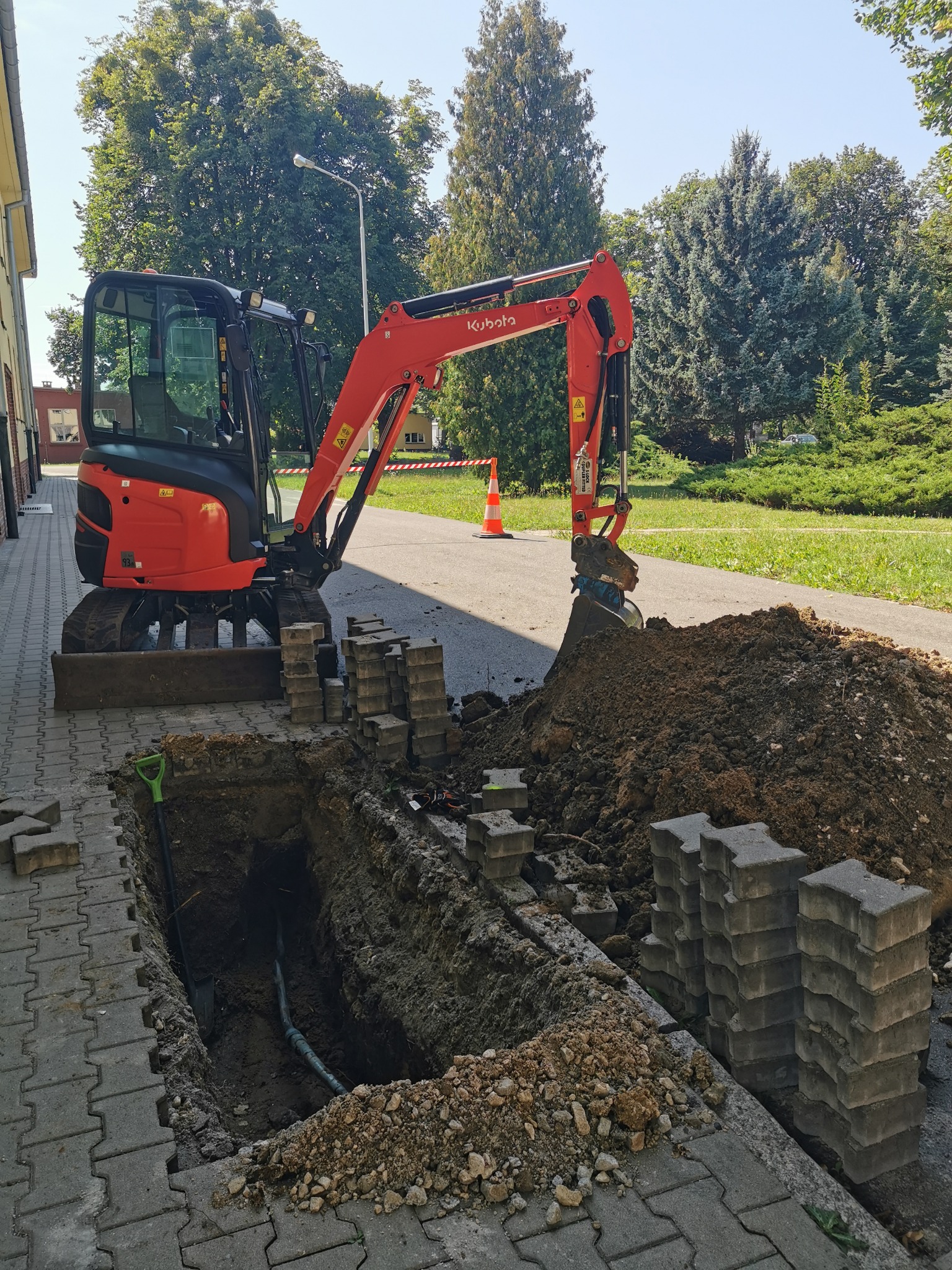 Czerwona mini koparka Kubota przy wykopie w chodniku z kostki, widoczne ułożone kostki, wykopana ziemia i rury w wykopie, na drugim planie zieleń i budynek.