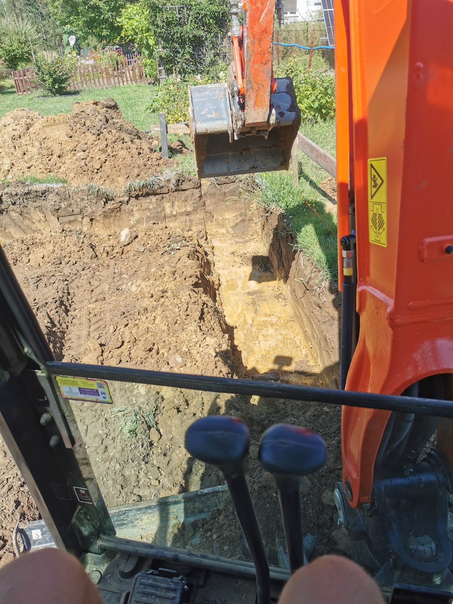 Widok z kabiny koparki na wykop pod fundamenty, z widoczną stertą ziemi i fragmentem krajobrazu z ogrodem w tle.