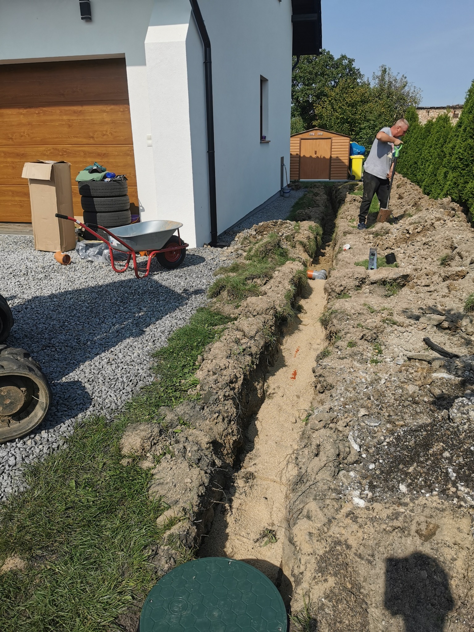 Pracownik kopie wąski wykop w ziemi obok domu, widoczne rury kanalizacyjne, taczka, opony i narzędzia