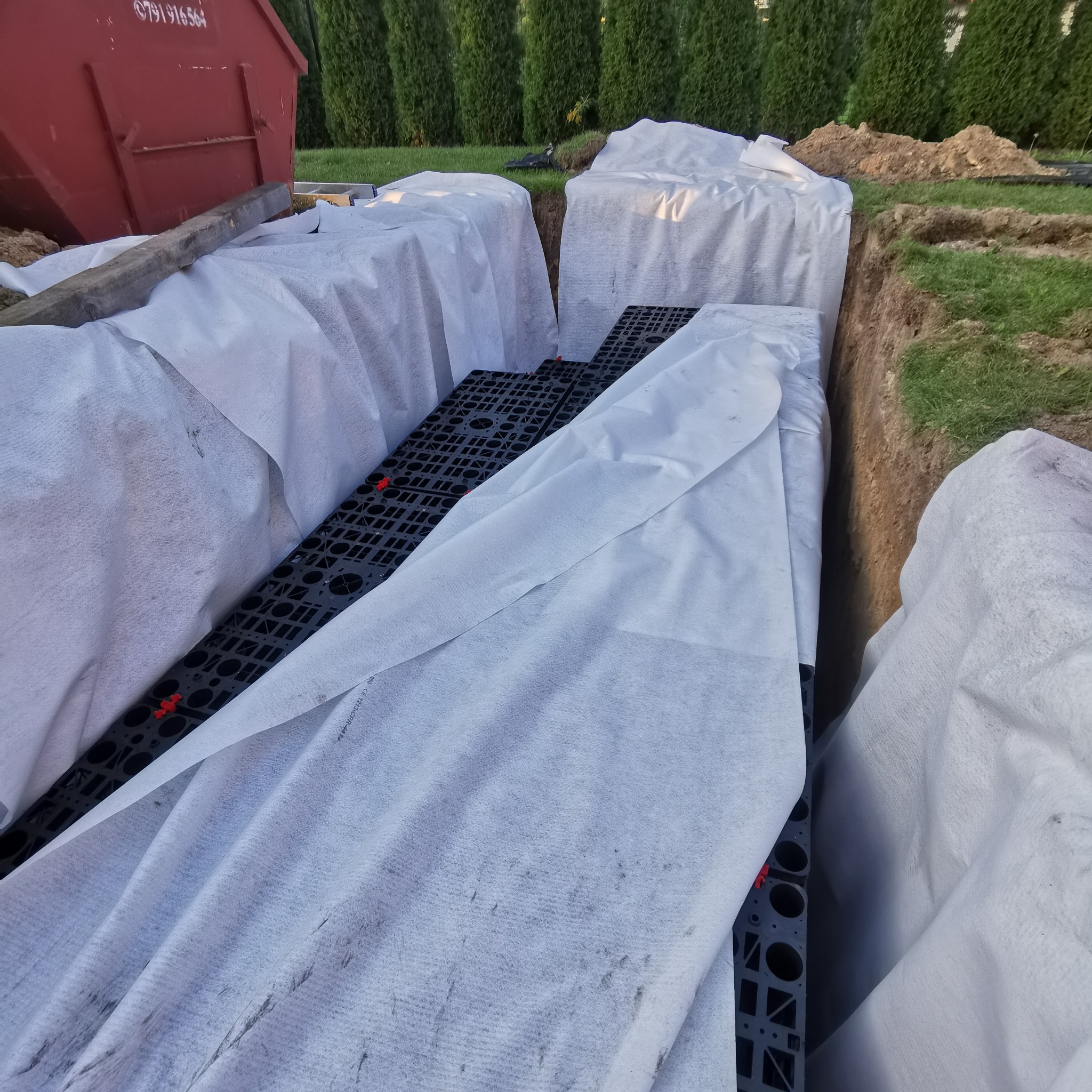 Wykop w ziemi z ułożonymi elementami drenażowymi, przykrytymi białą geowłókniną, widoczny kontener na gruz i trawnik w tle.