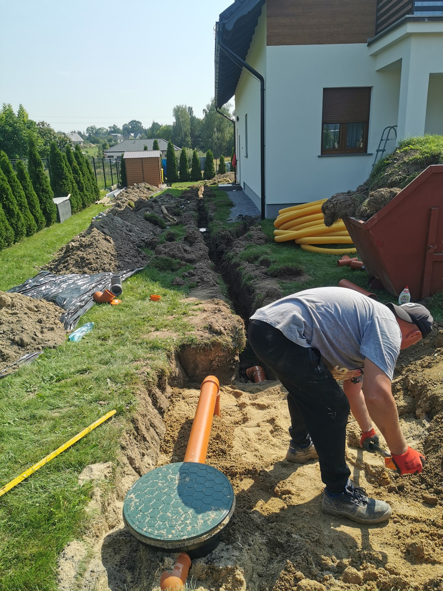 Pracownik w czerwonych rękawicach kopie łopatą w wykopie pod drenaż rozsączający, widoczna studzienka drenażowa z zieloną pokrywą, rury drenażowe, dom jednorodzinny w tle.