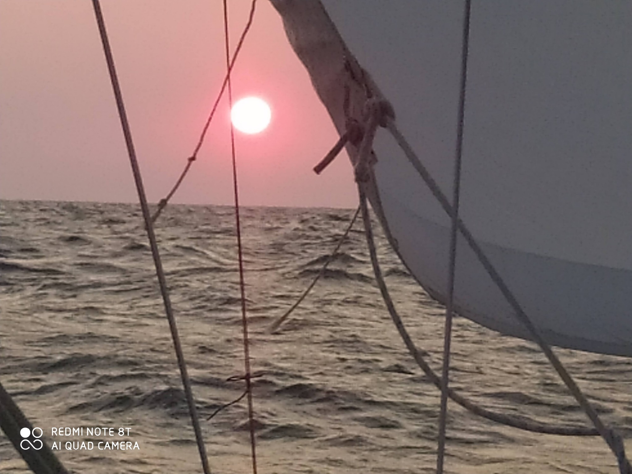 Widok z żaglówki na zachodzące słońce nad wzburzonym morzem, z fragmentem żagla i lin na pierwszym planie.