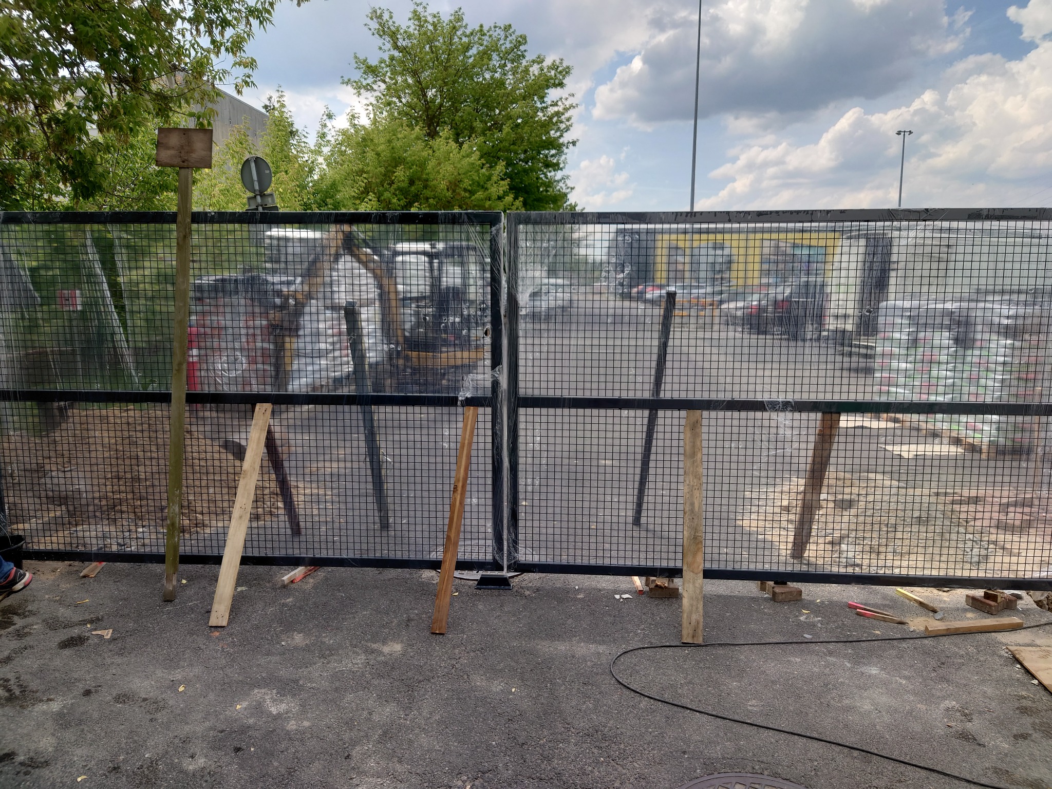 Tymczasowe ogrodzenie z siatki metalowej zabezpieczające teren budowy, widoczne elementy wsparcia z drewna, w tle zaparkowane samochody i sprzęt budowlany.