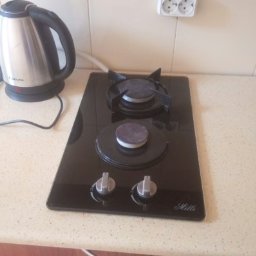 Natali Cleaning - Czarna, dwupalnikowa płyta gazowa marki Hilli, zamontowana w jasnym blacie kuchennym, obok czajnika elektrycznego i gniazdka. Palniki przykryte, pokrętła w pozycji wyłączonej.