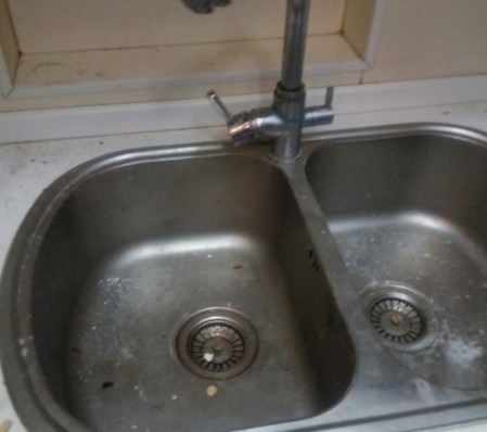Brudny, dwukomorowy zlewozmywak ze stali nierdzewnej z osadem i resztkami, widoczne niedomyte fragmenty na powierzchni i w odpływach.