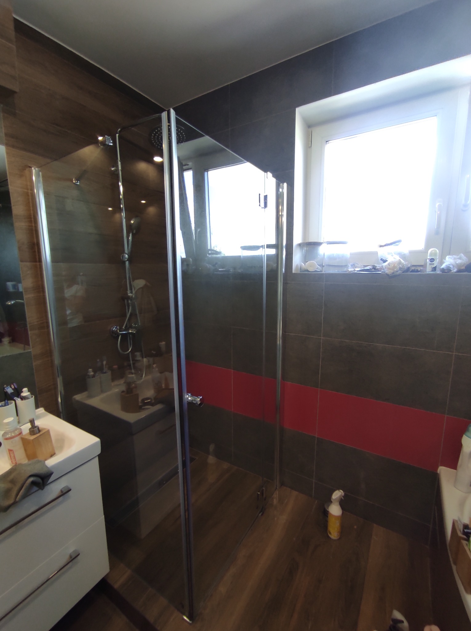 Nowoczesna kabina prysznicowa z transparentnego szkła w łazience z brązowo-czerwonymi płytkami, widoczna umywalka z szafką i okno z akcesoriami.