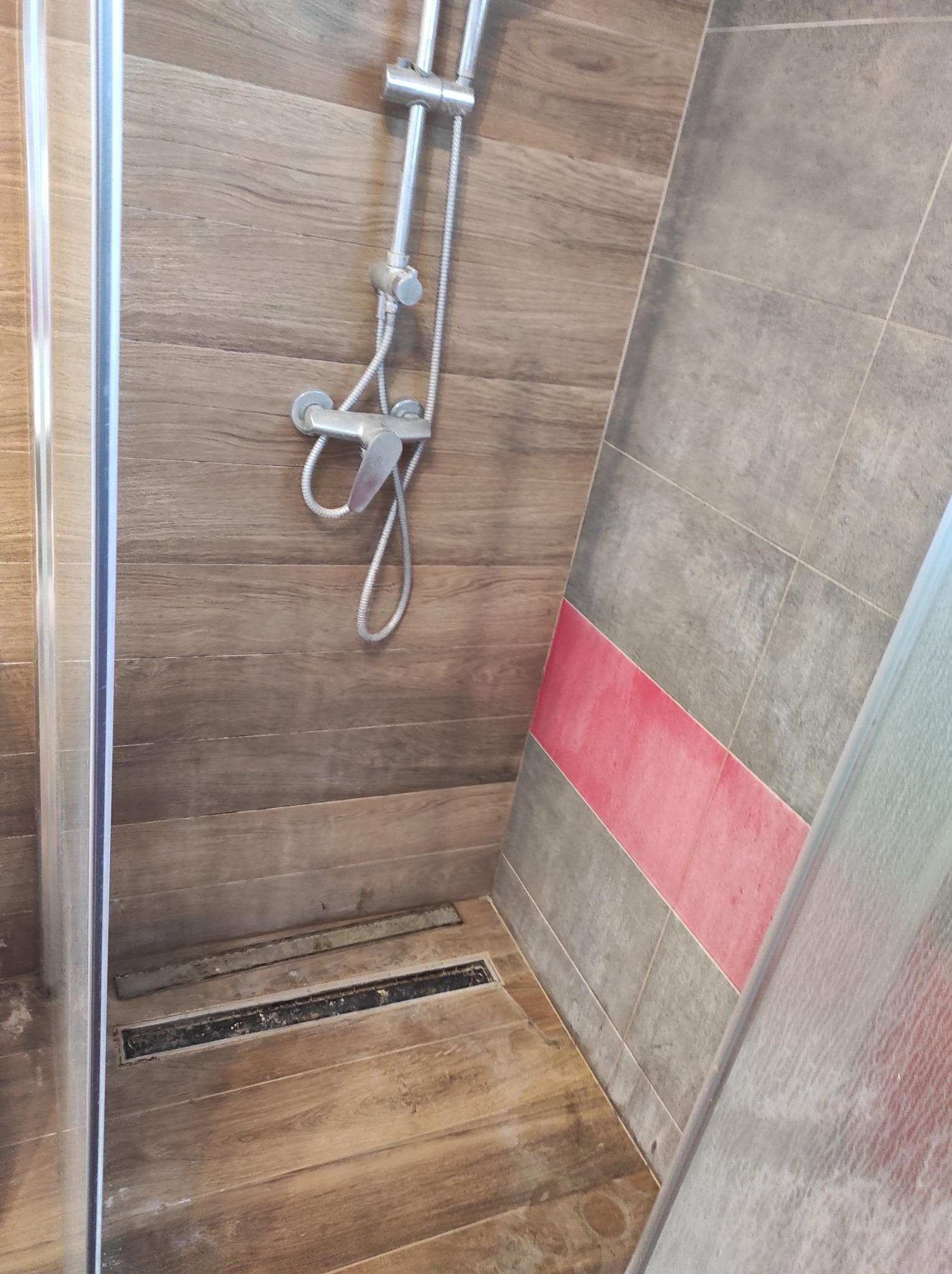 Wnętrze kabiny prysznicowej z odpływem liniowym, brązowe płytki imitujące drewno na ścianach i podłodze, szare płytki z różowym pasem na bocznej ścianie, widoczna bateria prysznicowa z deszczownicą.