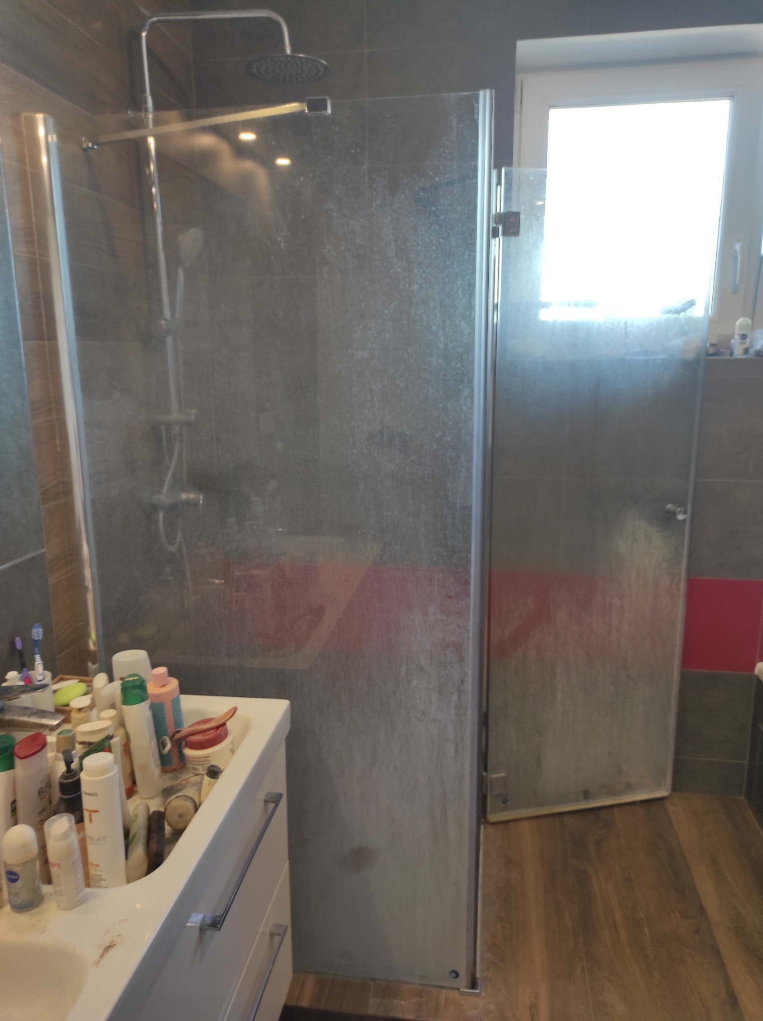 Zabrudzona kabina prysznicowa ze śladami po wodzie, widok na nieuporządkowaną umywalkę z kosmetykami i fragment okna z jasnym światłem.