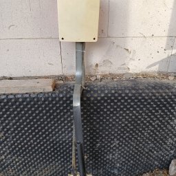 elmaro - Biała puszka elektryczna zamontowana na surowej ścianie z betonu komórkowego, z widocznymi przewodami w szarych peszlach, wychodzącymi z ziemi i przymocowanymi do ściany, z czarną folią...