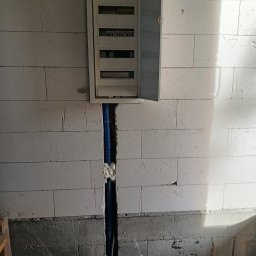 elmaro - Otwarta skrzynka rozdzielcza z widocznymi bezpiecznikami na ścianie z betonu komórkowego, z niebieskim peszlem w bruździe wypełnionej pianką montażową.