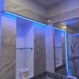 elmaro - Łazienka z podświetlanymi wnękami wykończonymi płytkami imitującymi marmur, widoczna szklana półka z papierem toaletowym i odbicie kabiny prysznicowej w lustrze.