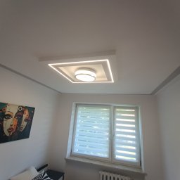 elmaro - Nowoczesne oświetlenie sufitowe LED w kształcie kwadratu z okrągłym kloszem w centrum, zamontowane w białym suficie w pokoju z obrazem na ścianie i widokiem na okno z żaluzjami.