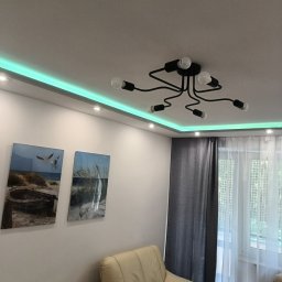 elmaro - Nowoczesny salon z podświetleniem LED na suficie, czarną lampą sufitową z odsłoniętymi żarówkami, dwoma obrazami na ścianie i beżowym fotelem.
