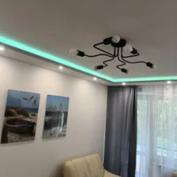 Nowoczesny salon z podświetleniem LED na suficie, czarną lampą sufitową z odsłoniętymi żarówkami, dwoma obrazami na ścianie i beżowym fotelem.