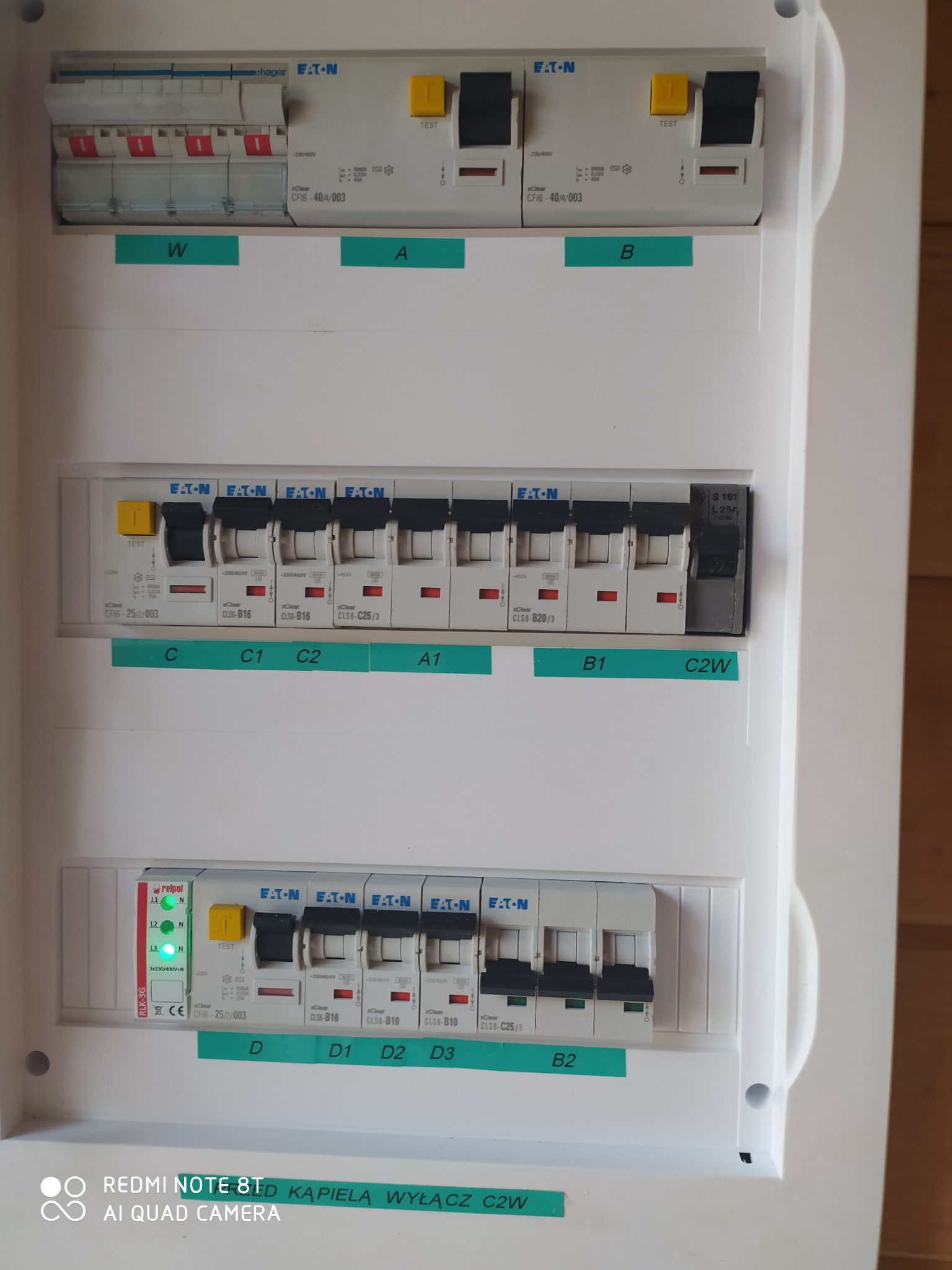 Tablica rozdzielcza z bezpiecznikami Eaton i Hager, opisana zielonymi etykietami z oznaczeniami obwodów, widoczne różne typy wyłączników i przekaźnik Relpol RLK-3G.