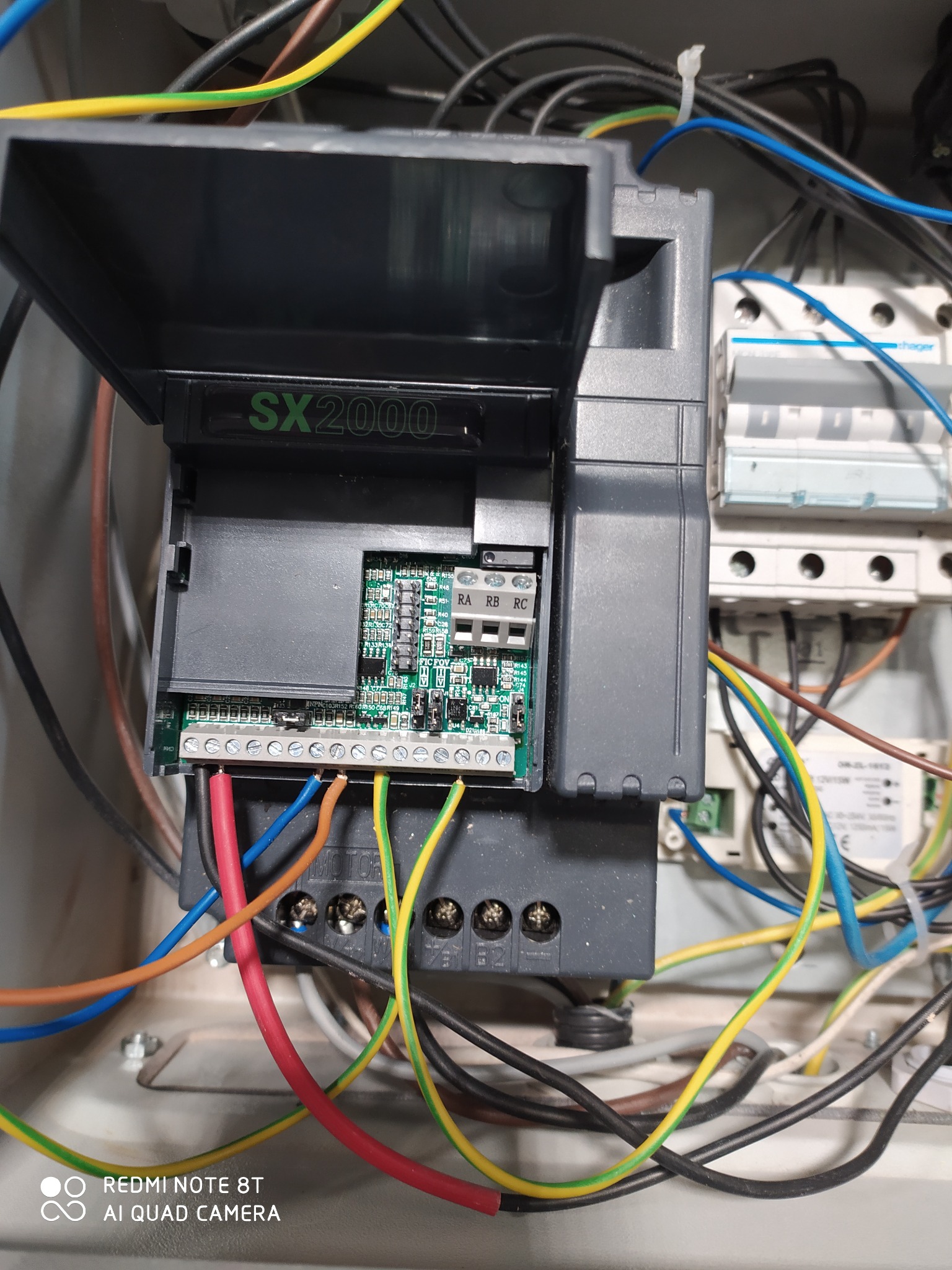 Otwarta obudowa sterownika SX2000 z widoczną płytką drukowaną i podłączonymi przewodami w kolorach czerwonym, niebieskim, brązowym i żółto-zielonym, zamontowanego w skrzynce elektrycznej obok...