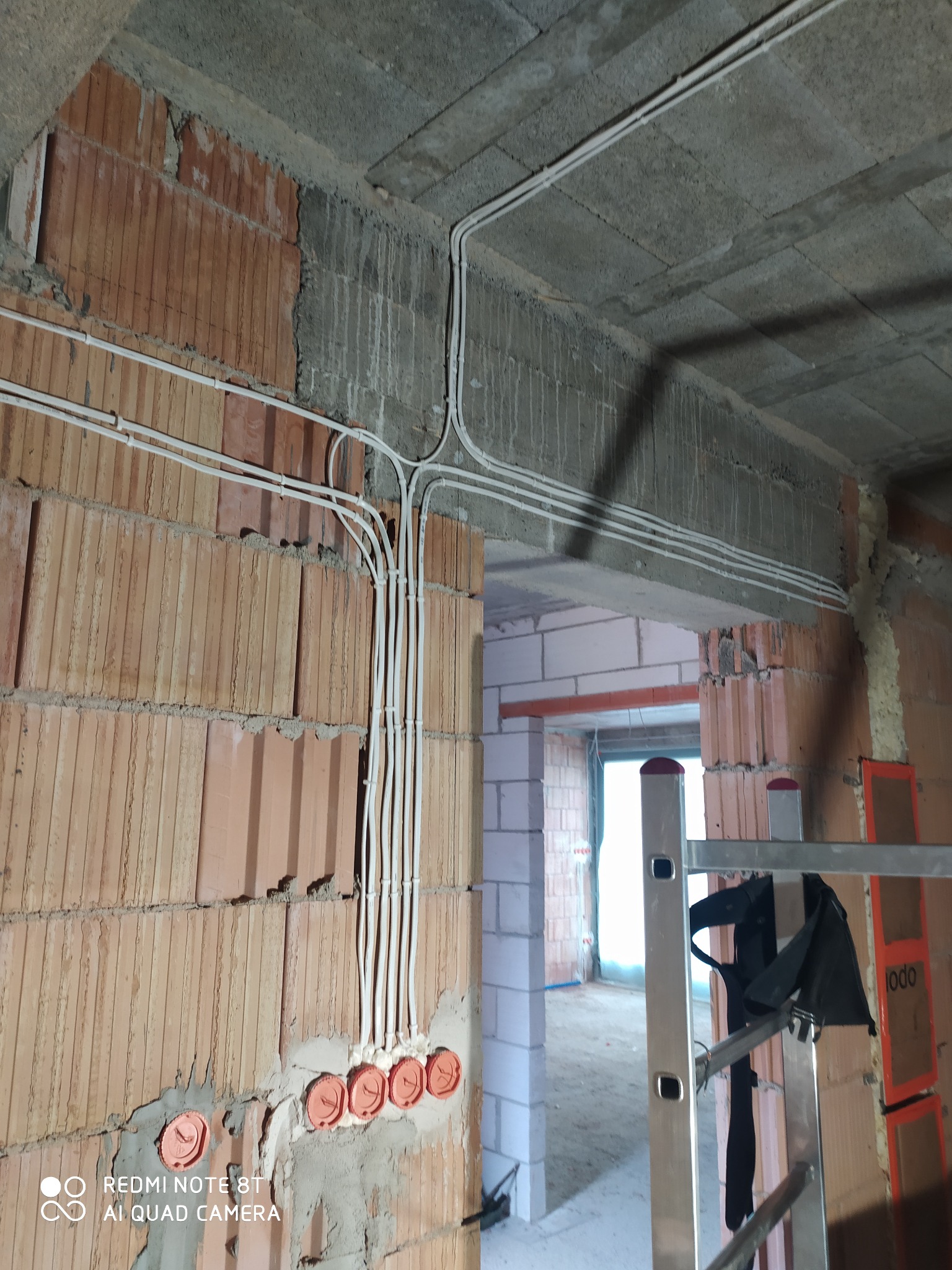 Surowe wnętrze w trakcie prac: widoczne kable elektryczne prowadzone po ceglanej ścianie i betonowym suficie, puszki instalacyjne w ścianie, w tle fragment drabiny i kolejne pomieszczenie w stanie...