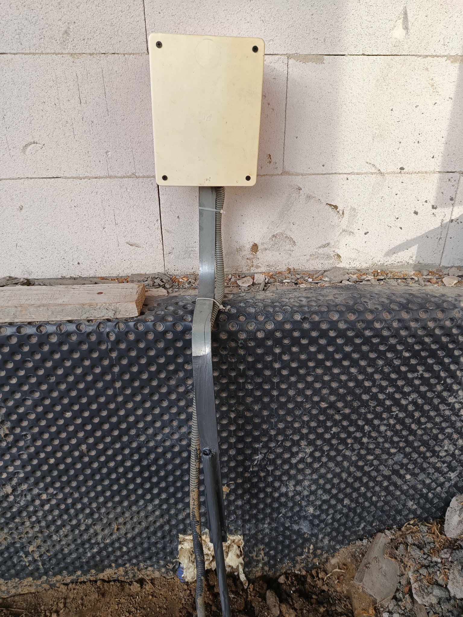 Biała puszka elektryczna zamontowana na surowej ścianie z betonu komórkowego, z widocznymi przewodami w szarych peszlach, wychodzącymi z ziemi i przymocowanymi do ściany, z czarną folią...
