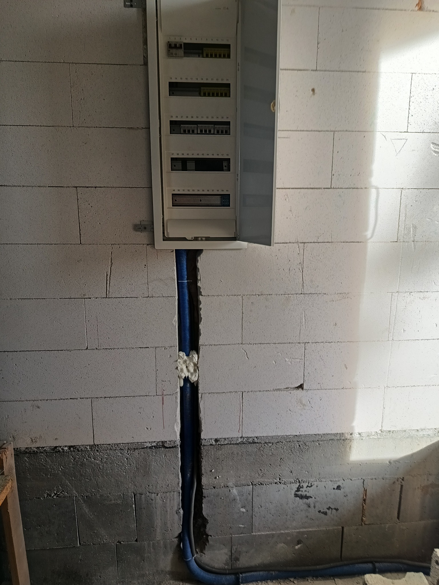 Otwarta skrzynka rozdzielcza z widocznymi bezpiecznikami na ścianie z betonu komórkowego, z niebieskim peszlem w bruździe wypełnionej pianką montażową.