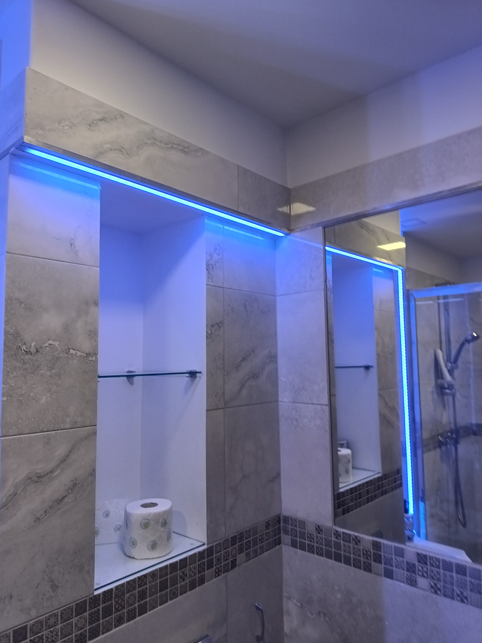 Łazienka z podświetlanymi wnękami wykończonymi płytkami imitującymi marmur, widoczna szklana półka z papierem toaletowym i odbicie kabiny prysznicowej w lustrze.