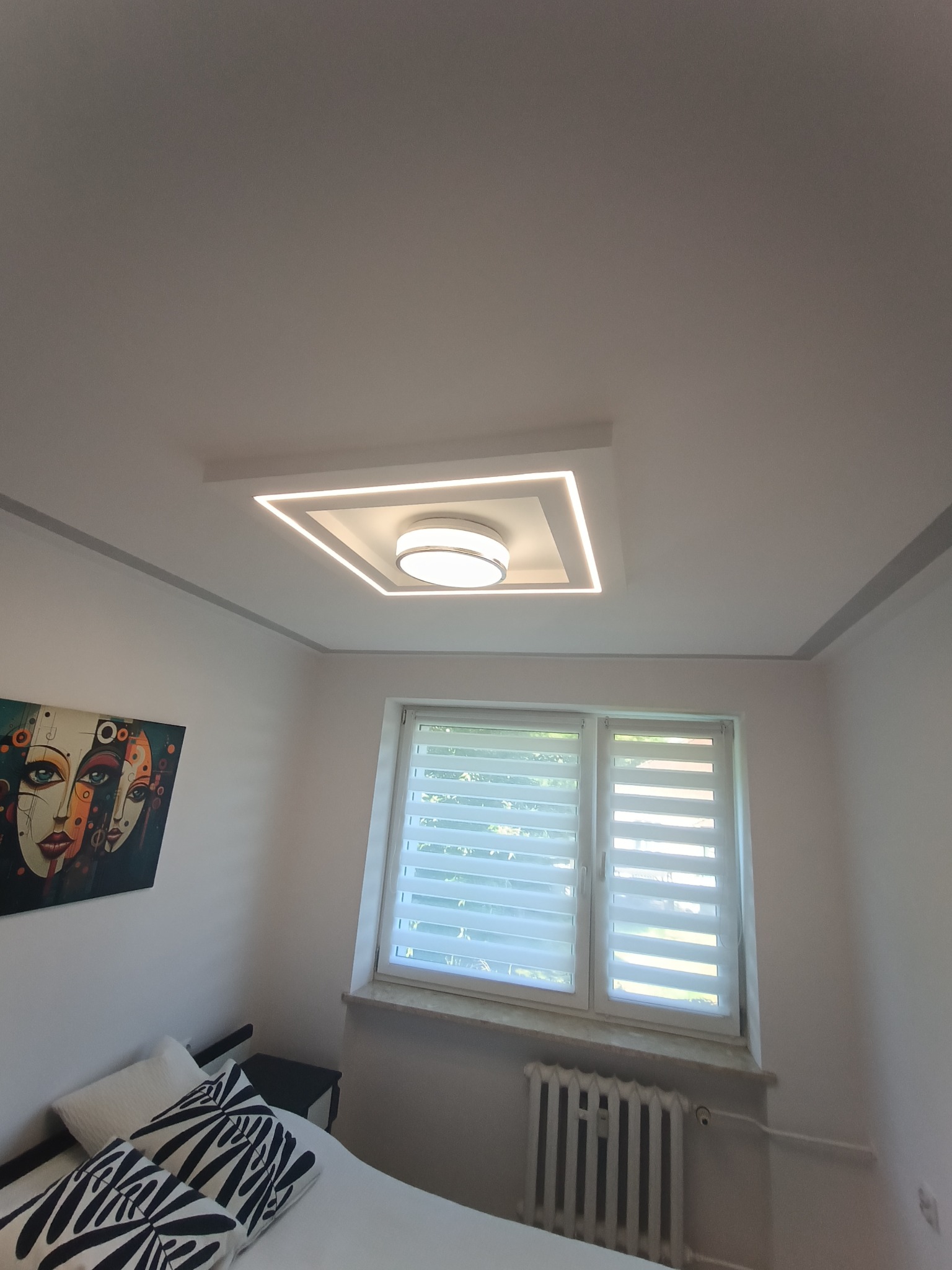 Nowoczesne oświetlenie sufitowe LED w kształcie kwadratu z okrągłym kloszem w centrum, zamontowane w białym suficie w pokoju z obrazem na ścianie i widokiem na okno z żaluzjami.