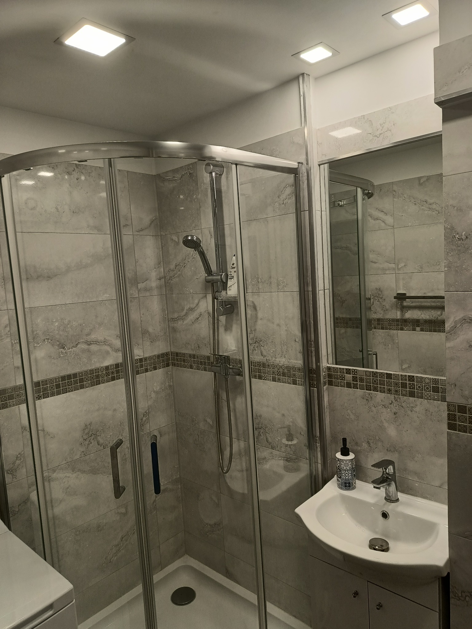 Wykończona łazienka z kabiną prysznicową z zaokrąglonymi drzwiami, umywalką w szafce i szarymi płytkami imitującymi marmur z dekoracyjnym pasem na ścianach.