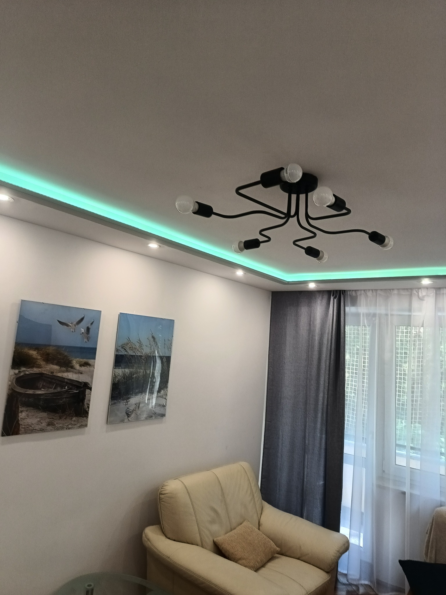 Nowoczesny salon z podświetleniem LED na suficie, czarną lampą sufitową z odsłoniętymi żarówkami, dwoma obrazami na ścianie i beżowym fotelem.