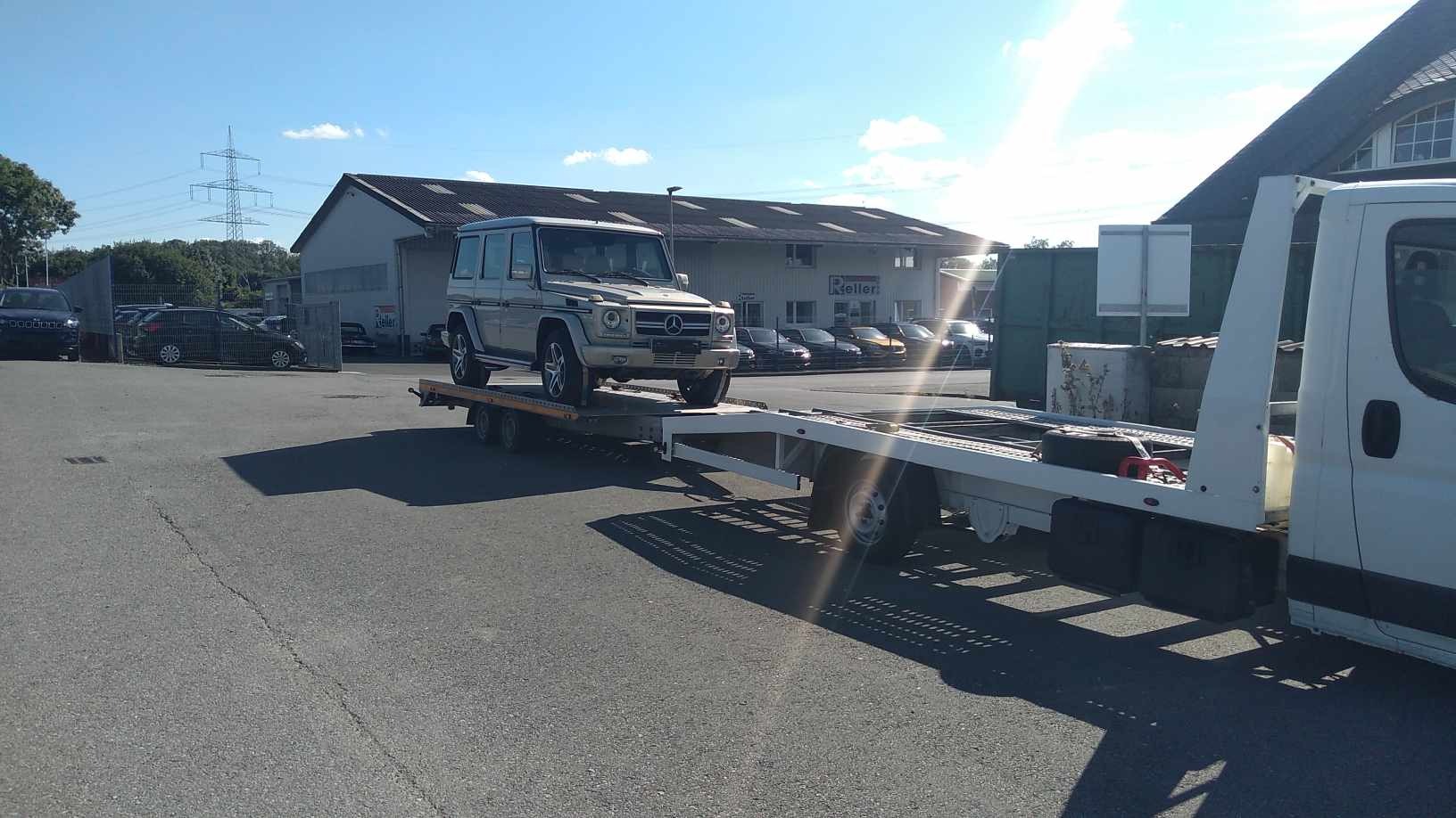 Srebrny Mercedes G-klasy na platformie lawety, na tle parkingu samochodowego i budynku firmy Keller w słoneczny dzień.
