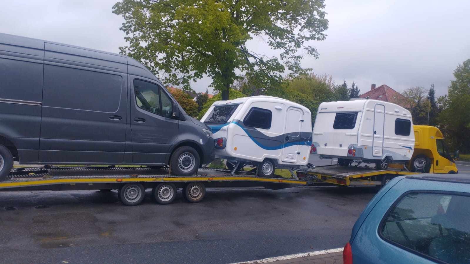 Laweta przewozi trzy małe białe przyczepy kempingowe z niebieskimi i szarymi akcentami; pierwsza przyczepa częściowo zasłonięta przez szarego busa, ostatnia przez żółty pojazd transportowy.