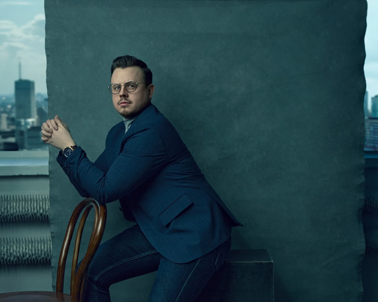 Portret mężczyzny w okularach i granatowym garniturze, opierającego się o krzesło na tle abstrakcyjnego tła z widokiem na miasto.