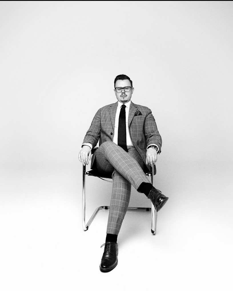 Portret adwokata w kraciastej marynarce i spodniach, siedzącego na chromowanym krześle na białym tle, czarno-białe zdjęcie.