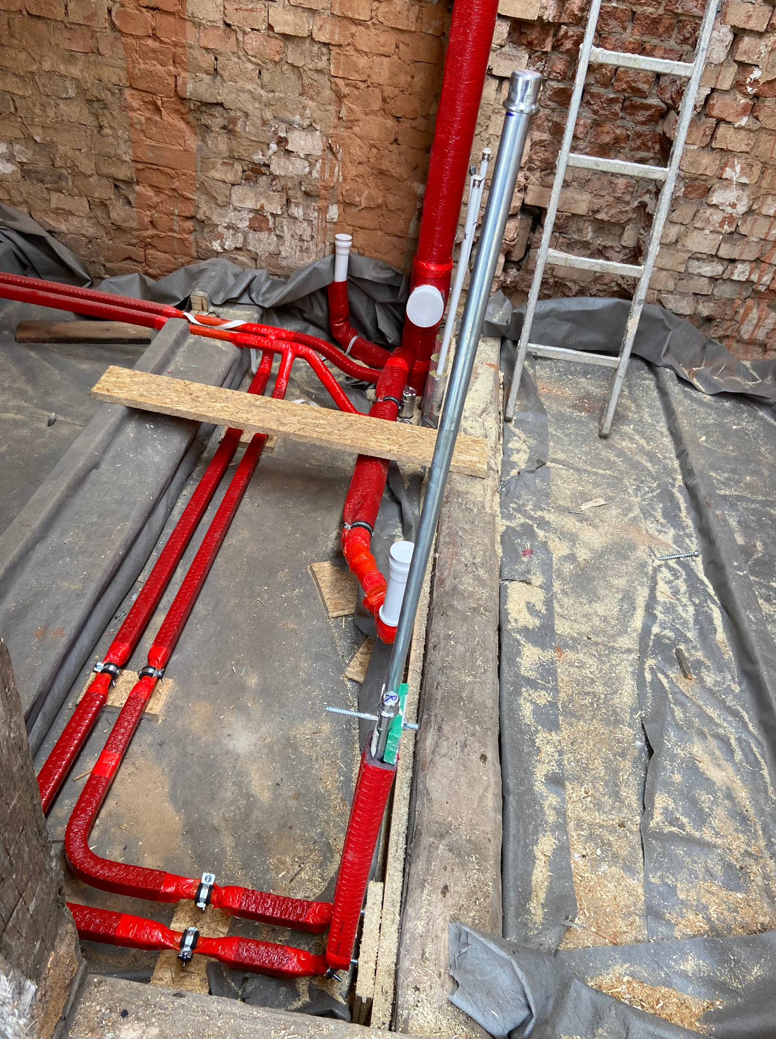 Czerwone rury instalacji grzewczej, częściowo owinięte izolacją, zamontowane w podłodze w trakcie prac remontowych, widoczna drabina i ceglana ściana w tle.