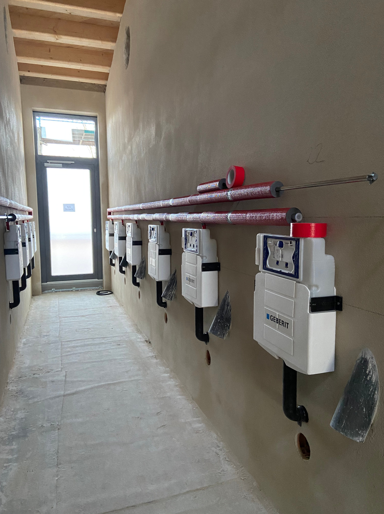 Rząd białych podtynkowych stelaży WC marki Geberit zamontowanych na surowej ścianie w trakcie instalacji, widoczne rury w izolacji z czerwoną taśmą, surowy beton na podłodze i okno w tle.