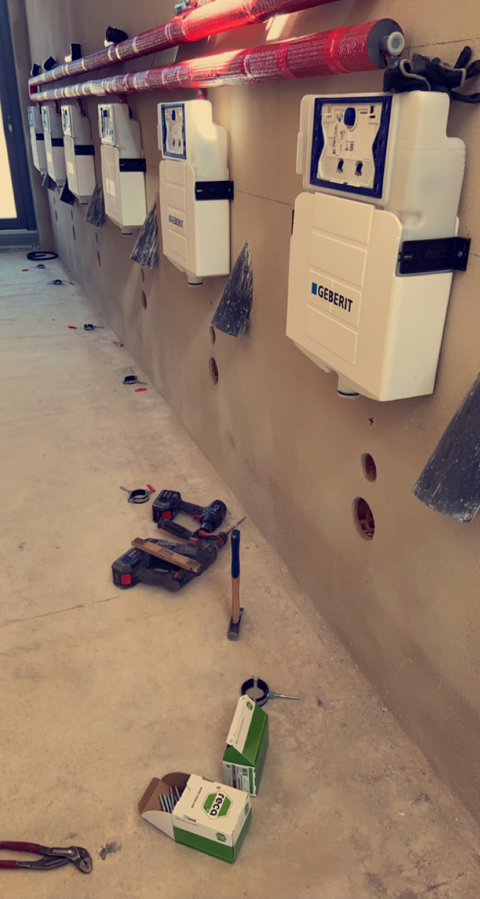 Montaż podtynkowych stelaży WC marki Geberit, widoczne narzędzia i materiały montażowe, rury instalacji sanitarnej pod sufitem, surowy stan wykończenia wnętrza.