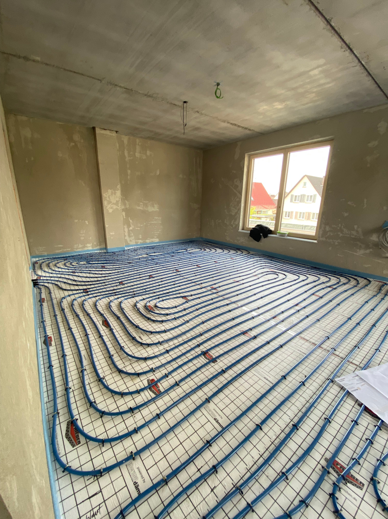 Niedokończona instalacja ogrzewania podłogowego z niebieskimi rurkami na białej macie z czarną siatką w surowym pokoju z oknem.