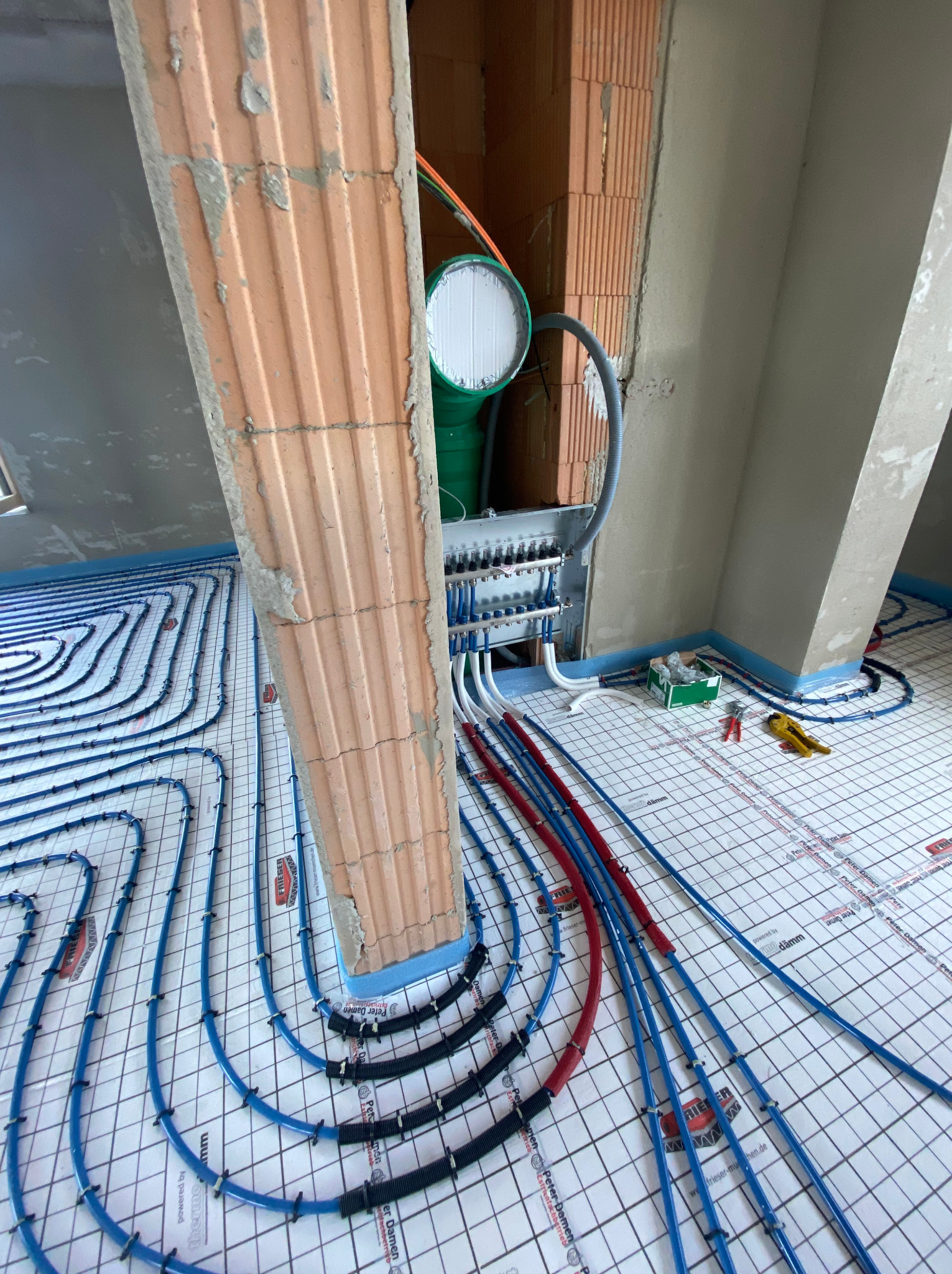 Rozłożone rury grzewcze w systemie podłogowym, widoczny rozdzielacz, narzędzia i surowy mur z cegły dziurawki w tle.