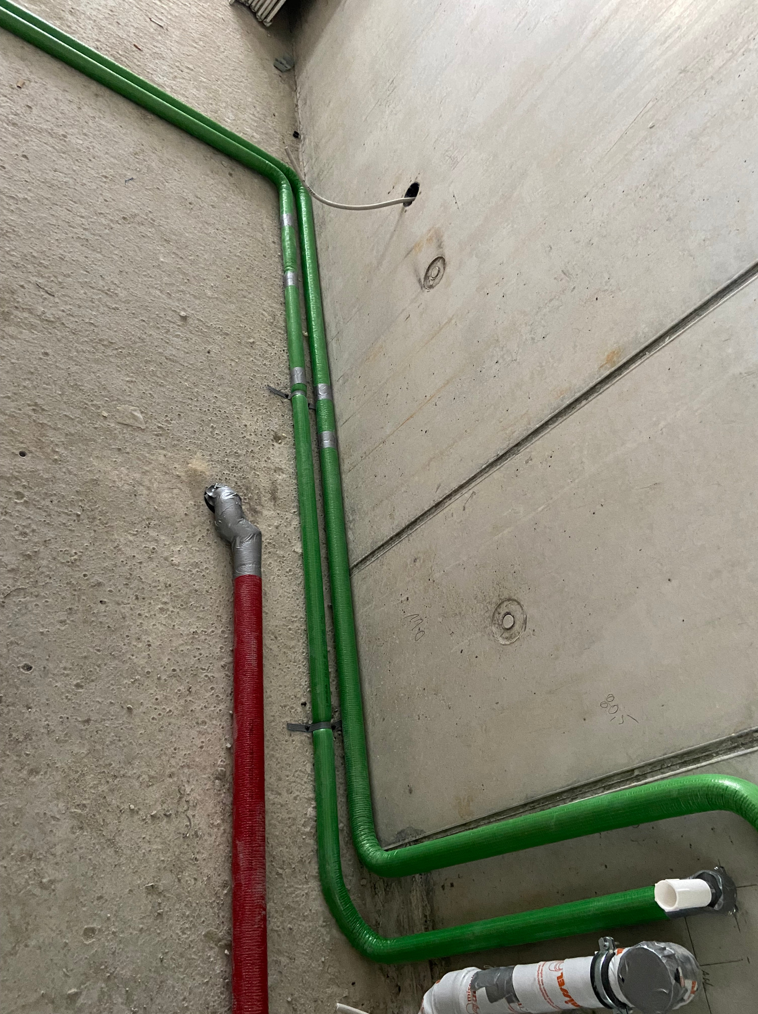 Surowe wnętrze z betonowymi ścianami, gdzie widoczne są pionowe rury grzewcze w kolorze zielonym, przymocowane klamrami, oraz rura czerwona z prowizorycznym uszczelnieniem z taśmy srebrnej.